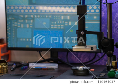 A microscope is positioned on a workstation next to a computer screen displaying a circuit board design. Tools are scattered. A microscope is positioned on a workstation next to a computer screen displaying a circuit board design. Tools are scattered. 140461037