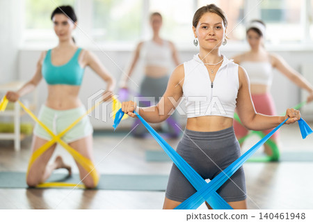 Young woman performs exercises with resistance band during Pilates strength training, kneeling Young woman performs exercises with resistance band during Pilates strength training, kneeling 140461948