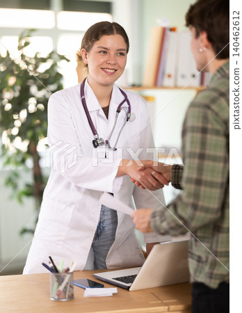 Smiling young female doctor or nurse wearing white uniform with stethoscope shaking hands with young guy patient congratulating on good test results or recovery from illness in medical office at Smiling young female doctor or nurse wearing white uniform with stethoscope shaking hands with young guy patient congratulating on good test results or recovery from illness in medical office at 140462612