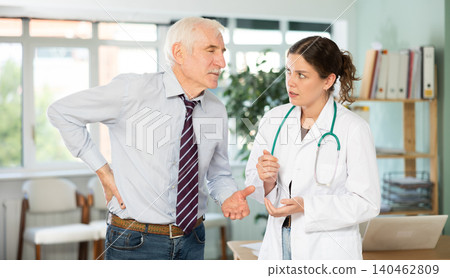 Senior man holds sore spot with his hand, complains about lower back pain to girl doctor Senior man holds sore spot with his hand, complains about lower back pain to girl doctor 140462809