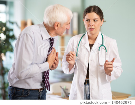 Senior man holds sore spot with his hand, complains about pain in heart to girl doctor, Senior man holds sore spot with his hand, complains about pain in heart to girl doctor, 140462975
