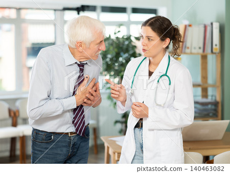 Elderly man patient complaining of pain to young woman doctor or nurse wearing white medical coat with stethoscope standing in office of medical center Elderly man patient complaining of pain to young woman doctor or nurse wearing white medical coat with stethoscope standing in office of medical center 140463082