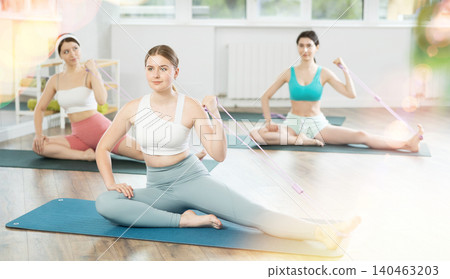 Young woman performs stretches elastic band with hand and foot while sitting on mat. Young woman performs stretches elastic band with hand and foot while sitting on mat. 140463203