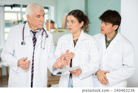 Three professional male and female doctors in white medical coat discussing and explaining results of medical test in doctor office in clinic Three professional male and female doctors in white medical coat discussing and explaining results of medical test in doctor office in clinic 140463236