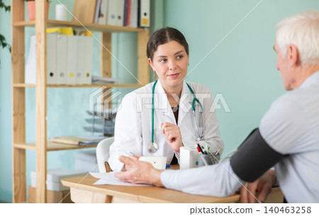 Experienced and polite young woman doctor or nurse in white uniform sitting at table and performing medical procedures to measure blood pressure elderly man patient in medical office at hospital 140463258