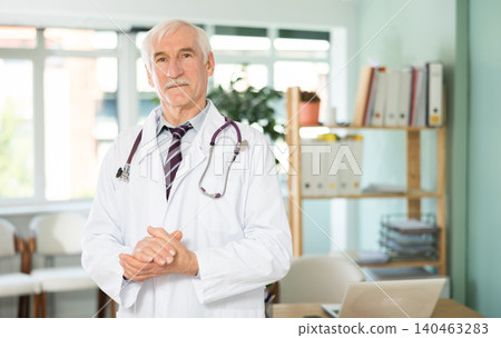Portrait of friendly elderly man doctor wearing white uniform standing with stethoscope in clinic Portrait of friendly elderly man doctor wearing white uniform standing with stethoscope in clinic 140463283