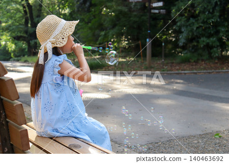 一個小女孩在公園長椅上玩肥皂泡,玩得不亦樂乎。 一個小女孩在公園長椅上玩肥皂泡,玩得不亦樂乎。 140463692