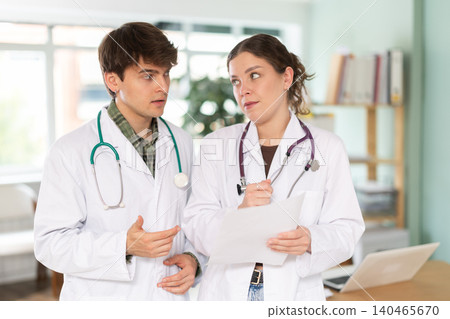 Two professional young male and female doctors in white medical coat discussing and explaining results of medical test in doctor office in clinic Two professional young male and female doctors in white medical coat discussing and explaining results of medical test in doctor office in clinic 140465670