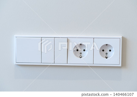 Two electrical sockets and three switches are mounted on a plain wall in a modern indoor setting. Two electrical sockets and three switches are mounted on a plain wall in a modern indoor setting. 140466107