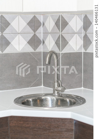 A corner sink setup features a simple faucet and gray tiled walls, suitable for kitchen tasks and food preparation. A corner sink setup features a simple faucet and gray tiled walls, suitable for kitchen tasks and food preparation. 140466131