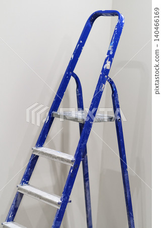 A ladder leans against a plain wall showing signs of use with paint drops on the floor below it after a home improvement task has taken place. 140466169