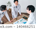 Three male office workers in work clothes are seen smiling and chatting while looking at documents in a conference room. 140466251