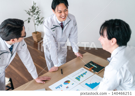 Three male office workers in work clothes are seen smiling and chatting while looking at documents in a conference room. 140466251