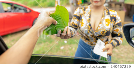 Local Thai customer reaches from car window to purchase fresh kratom leaves from traditional roadside vendor for recreational and medicinal use. Everyday commerce in rural Thailand's herbal market. Local Thai customer reaches from car window to purchase fresh kratom leaves from traditional roadside vendor for recreational and medicinal use. Everyday commerce in rural Thailand's herbal market. 140467259