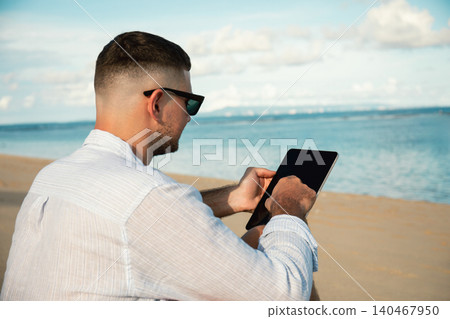 Man uses tablet while sitting on sandy beach by ocean 140467950