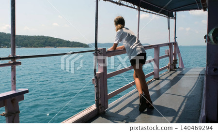 Teenage girl standing and leaning on ferry deck railing watching a tropical island scenery passing by during sea travel. Island journey concept Teenage girl standing and leaning on ferry deck railing watching a tropical island scenery passing by during sea travel. Island journey concept 140469294