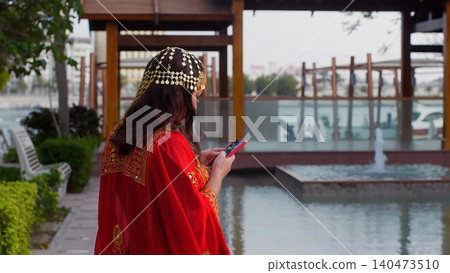 Woman wearing traditional red jalabiya using smartphone by canal Woman wearing traditional red jalabiya using smartphone by canal 140473510