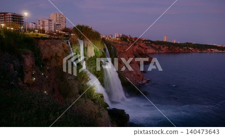 Waterfall cascading off coastal cliff at twilight with city lights 140473643
