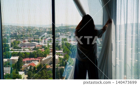 Woman opening curtains at high rise window overlooking city landscape Woman opening curtains at high rise window overlooking city landscape 140473699