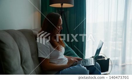 Woman working on laptop while sitting on couch by window Woman working on laptop while sitting on couch by window 140473701