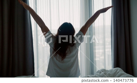 Woman stretching arms wide by window in morning bedroom Woman stretching arms wide by window in morning bedroom 140473776