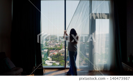 Woman opening sheer curtains at high rise window overlooking cityscape Woman opening sheer curtains at high rise window overlooking cityscape 140473987