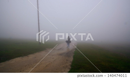 Person walking solitary dirt road through dense fog, rural landscape Person walking solitary dirt road through dense fog, rural landscape 140474011
