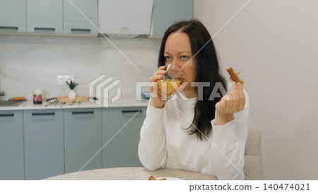 Woman drinking juice, eating bread in kitchen 140474021