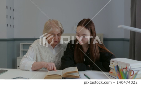 Senior woman mentoring teenage girl reading textbook together at desk Senior woman mentoring teenage girl reading textbook together at desk 140474022