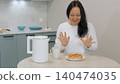 Woman enjoying hot croissant with electric kettle in kitchen 140474035