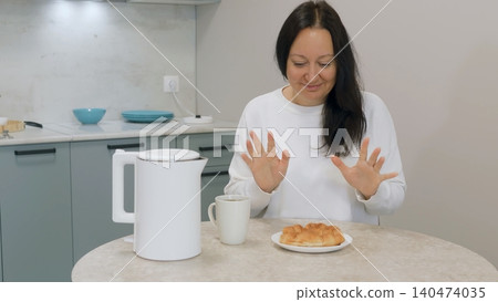 Woman enjoying hot croissant with electric kettle in kitchen 140474035