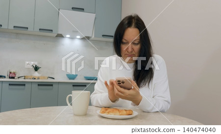 Woman using smartphone during breakfast in modern kitchen Woman using smartphone during breakfast in modern kitchen 140474040