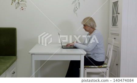 Senior woman typing on laptop at white desk in bright home office Senior woman typing on laptop at white desk in bright home office 140474084