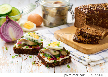 Black rye bread with butter, herring fish, boiled eggs, capers and red onion on a table. Traditional Danish open sandwich smorrebrod. Black rye bread with butter, herring fish, boiled eggs, capers and red onion on a table. Traditional Danish open sandwich smorrebrod. 140474418
