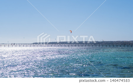 Long wooden pier stretches into the ocean, with a kite surfer visible in the background against a clear blue sky and shimmering water Long wooden pier stretches into the ocean, with a kite surfer visible in the background against a clear blue sky and shimmering water 140474583