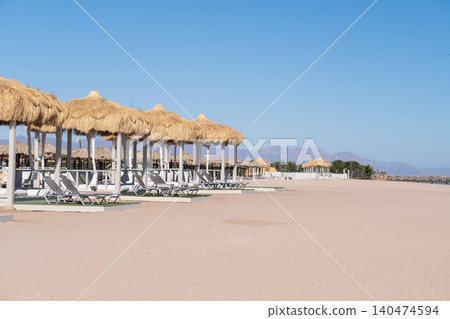 Beachfront scene featuring thatched-roof cabanas and lounge chairs on sandy shore, with a clear blue sky and distant mountains in the background Beachfront scene featuring thatched-roof cabanas and lounge chairs on sandy shore, with a clear blue sky and distant mountains in the background 140474594