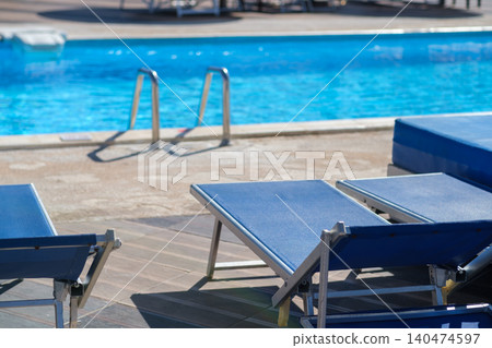Blue lounge chairs are arranged next to a clear swimming pool featuring steps leading into the water, with a sunlit patio area visible 140474597