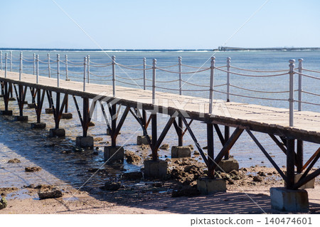 Wooden pier stretches over tranquil water with rocky shore visible below, featuring a clear blue sky and distant horizon in a coastal area 140474601