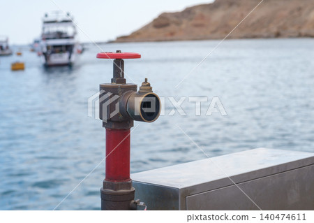 Red fire hydrant valve positioned on a dock with boats visible in the background and calm water reflecting the nearby shoreline 140474611