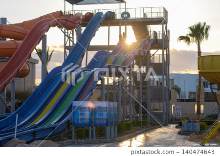 Colorful water slides are visible at an amusement park, with multiple levels and a sunset illuminating the scene against a clear sky 140474643