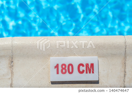Swimming pool edge features a depth marker labeled 180 cm, with a clear blue water surface shimmering in the background under sunlight Swimming pool edge features a depth marker labeled 180 cm, with a clear blue water surface shimmering in the background under sunlight 140474647