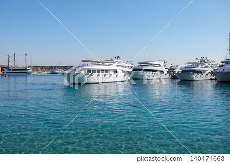 Luxury yachts are moored in clear blue water at a marina, with a bright sunny sky and distant boats visible in the background 140474668