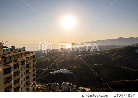 Beautiful golden sunset illuminates a coastal city, with buildings in the foreground and mountains rising in the distance along the serene water. Beautiful golden sunset illuminates a coastal city, with buildings in the foreground and mountains rising in the distance along the serene water. 140474837