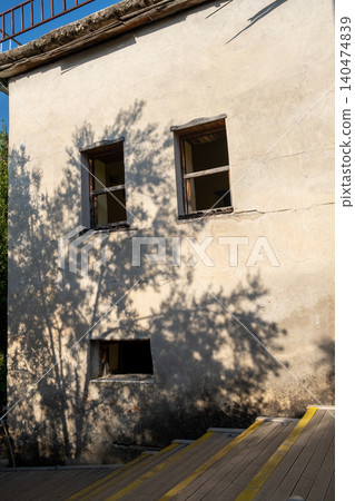 A sunny day reveals intricate shadows on an aged wall with windows, featuring a wooden ramp and greenery surrounding the structure. A sunny day reveals intricate shadows on an aged wall with windows, featuring a wooden ramp and greenery surrounding the structure. 140474839