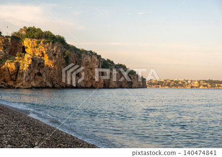 Cliffs rise above a calm sea as the sun sets, casting warm hues on the water, while pebbles line the beach in a serene coastal atmosphere. Cliffs rise above a calm sea as the sun sets, casting warm hues on the water, while pebbles line the beach in a serene coastal atmosphere. 140474841