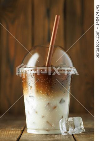 Iced latte coffee drink featuring distinct layers of espresso and milk with ice cubes in clear plastic cup on wooden surface 140474845
