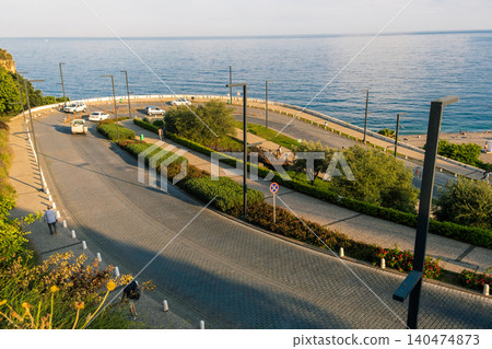 June 14, 2023, Antalya, Turkey. Vehicles park along a winding coastal road as people stroll near a park, enjoying the warm summer evening by the sea. June 14, 2023, Antalya, Turkey. Vehicles park along a winding coastal road as people stroll near a park, enjoying the warm summer evening by the sea. 140474873
