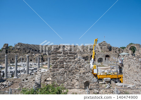 Restoration efforts are underway at an archaeological site featuring ancient ruins, historic columns, and stone structures against a bright blue sky. Restoration efforts are underway at an archaeological site featuring ancient ruins, historic columns, and stone structures against a bright blue sky. 140474900