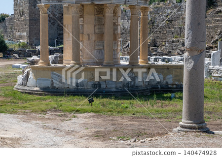 Ancient temple structure features distinct columns and stone remains, set within an archaeological site under a bright blue sky during midday. 140474928