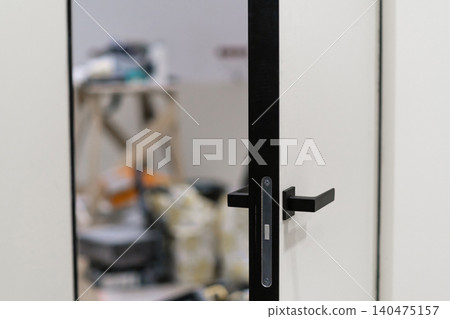 A modern door is shown in focus while tools and renovation supplies are visible in the background during a home renovation project. A modern door is shown in focus while tools and renovation supplies are visible in the background during a home renovation project. 140475157